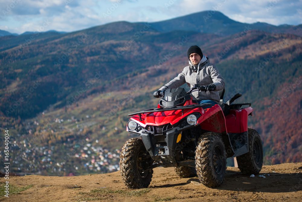 Guy in winter clothes on a red quad bike on a mountain top looking at the camera. Blurred background of mountain scenery Guy in winter clothes on a red quad bike on a mountain top looking at the camera. Blurred background of mountain scenery