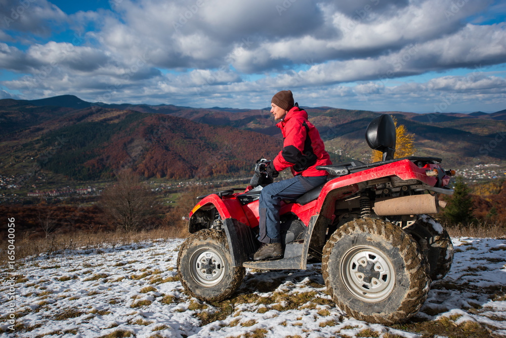 ATV quad bike with driver at the snowy slope against the blue cloudy sky, mountains and the town in the valley on a autumn sunny day in the highlands ATV quad bike with driver at the snowy slope against the blue cloudy sky, mountains and the town in the valley on a autumn sunny day in the highlands