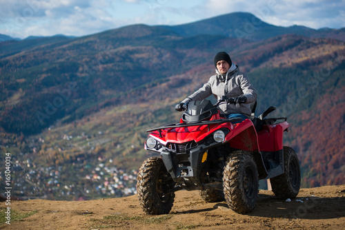 Guy in winter clothes on a red quad bike on a mountain top looking at the camera. Blurred background of mountain scenery