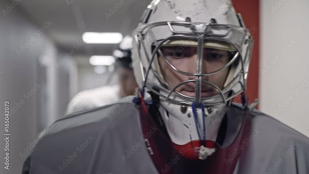 Dolly with close up of nervous hockey goaltender in full gear walking along hall towards ice rink before game; teammates following behind Dolly with close up of nervous hockey goaltender in full gear walking along hall towards ice rink before game; teammates following behind