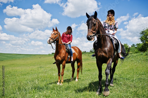 Tow young pretty girls riding a horses on a field at sunny day