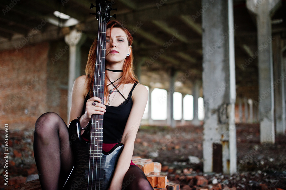 Red haired punk girl wear on black and red skirt, with bass guitar at abadoned place. Portrait of gothic woman musician.