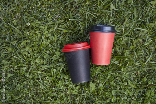 Two paper cups with coffee to take away, red and black cup on green grass outdoor breakfast