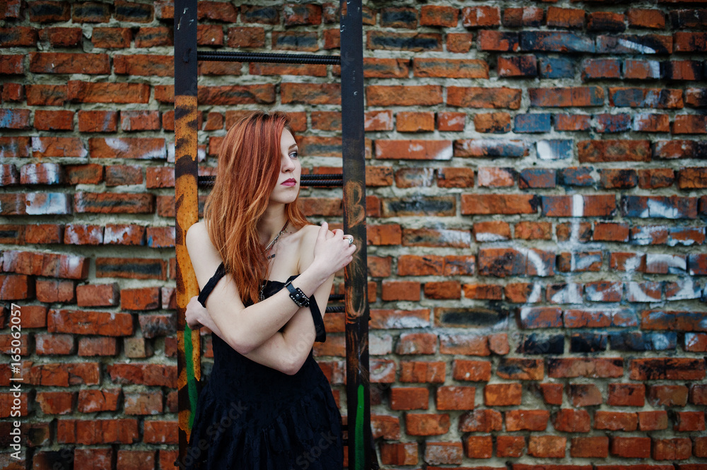 Red haired punk girl wear on black dress at the roof against brick wall with iron ladder.