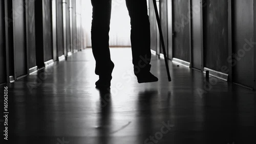 PAN with low-section of silhouette of legs of man with walking stick limping along dark hospital corridor