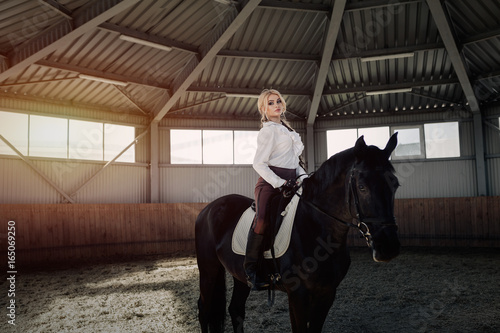 Beautiful elegant young blonde girl sits on a her black horse dressing uniform competition white blouse shirt and brown pants.