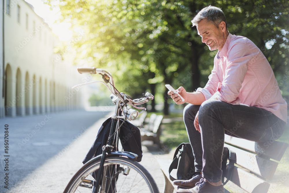 Smiling man with bicycle checking the phone on a park bench Smiling man with bicycle checking the phone on a park bench