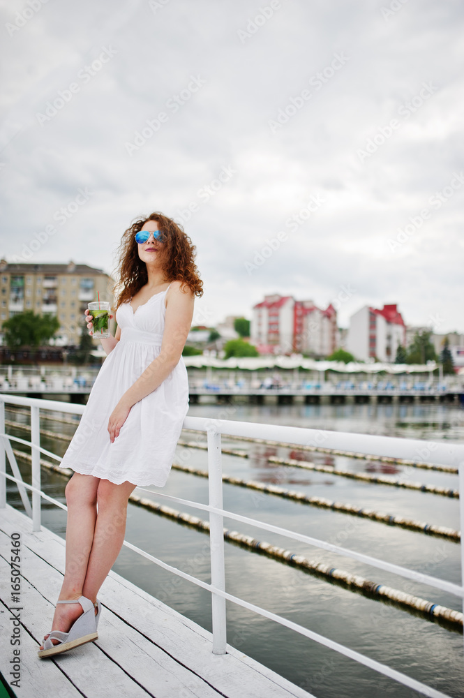 Portrait of an attractive young woman posing with her cocktail on a lakeside wearing sunglasses.