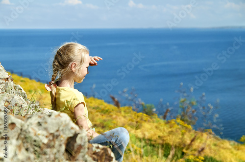 Nice blonde girl with pigtail sitting on the shore of a lake among the yellow flowers and looking into the distance holding her hand above the eyes 