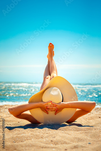 Back view of woman in hat lying and relaxing on beach with legs up.