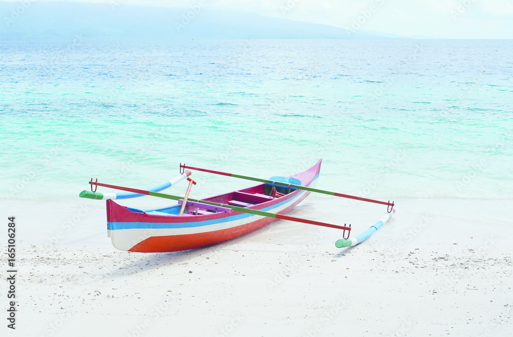 Red white blue colored small empty canoe boat stranded on a tropical white sand beach with blue ocean water on a sunny day in the afternoon.