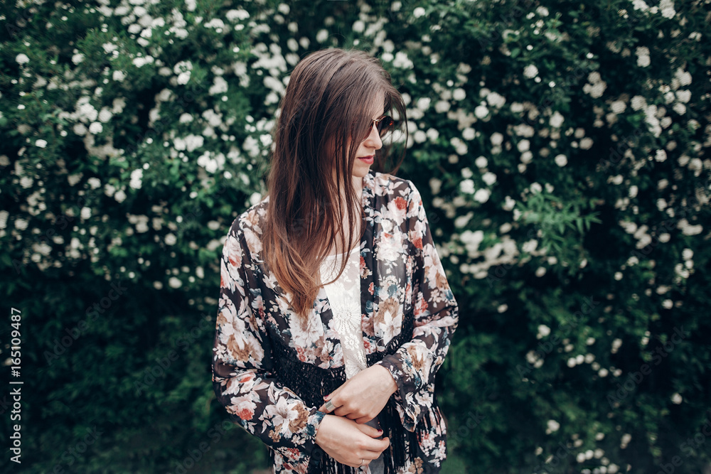 hipster woman in sunglasses posing on background of blooming bush with white flowers of spirea. boho girl sensual portrait in floral modern clothes in city street. space for text hipster woman in sunglasses posing on background of blooming bush with white flowers of spirea. boho girl sensual portrait in floral modern clothes in city street. space for text