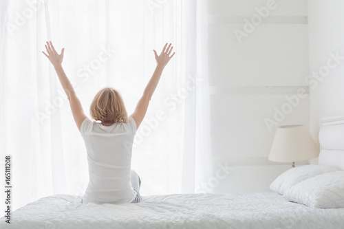 Happy woman sitting on bed in bedroom with hands raised up in the morning. Rear view