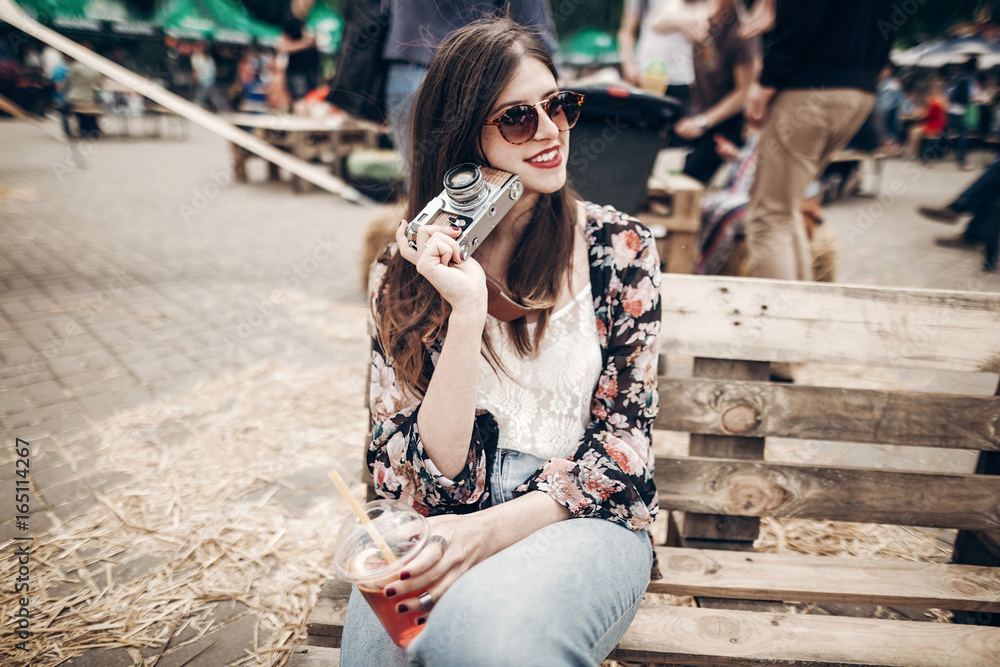 Fototapeta premium stylish hipster woman in sunglasses with red lips holding lemonade and old photo camera. boho girl holding cocktail and smiling at street food festival. summer vacation. space for text