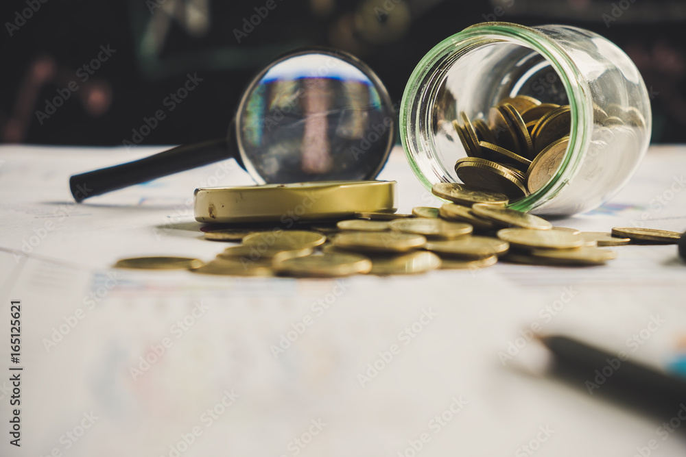 Coin in a bottle, magnifying glass, pen and calculator on financial chart and graph, accounting background Coin in a bottle, magnifying glass, pen and calculator on financial chart and graph, accounting background