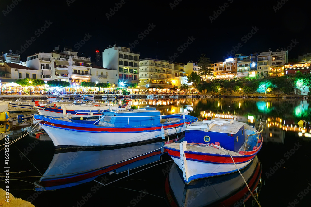 Fishing boats at para outra in night city of Crete, Agios Nikolaos in the background of seaside restaurants, cafes and hotels. Tourists relax in outdoor restaurant on the shores of lake Voulismeni