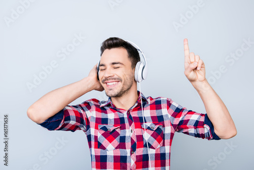 Close up portrait of cheerful young man enjoying listening to his favourite song at his holiday with closed eyes in big white modern earphones, wearing casual outfit