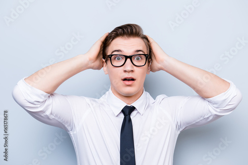 Oh wow! Really?! Young handsome student is shocked, holding his head, in a formal outfit and black stylish glasses, standing on pure background