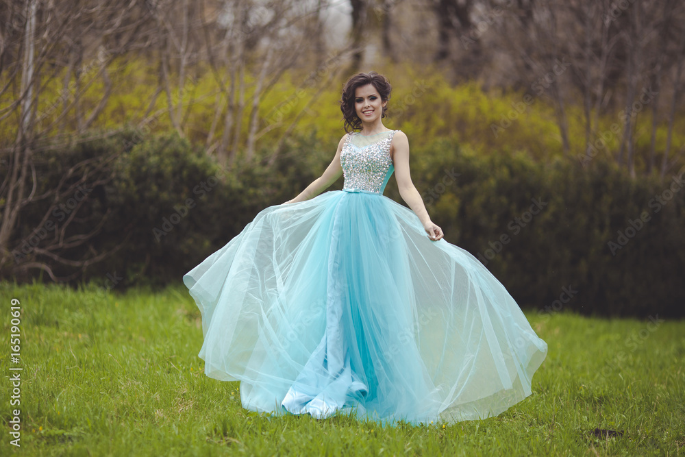 A beautiful graduate girl is spinning in a clearing in a blue fluffy dress. Elegant young woman in a beautiful dress in the park. Art photo.