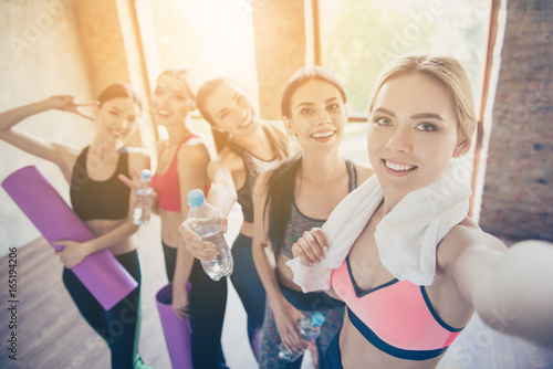 Selfie mania in gym! Five girlfriends in fashionable sport outfits are posing for a selfie photo, that blond is taking. They are all smiling, joyful after the workout