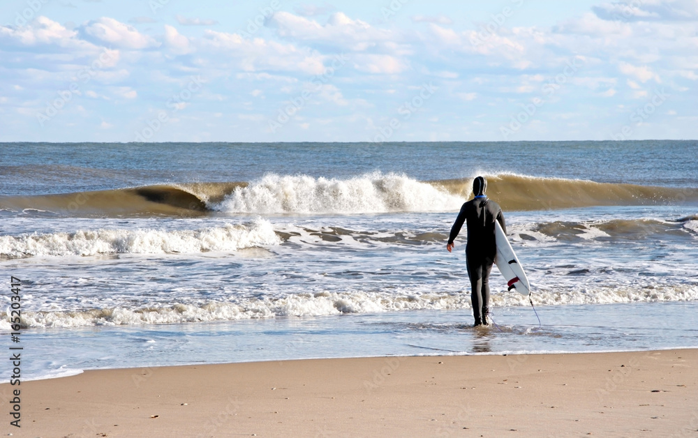 Surfer wearing wetsuit, carrying surfboard contemplating wave conditions.