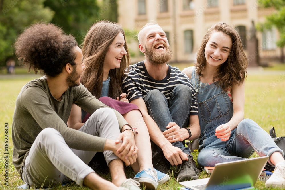 Happily laughing students on grass in University park. Group of young people sitting on lawn, having fun. Happily laughing students on grass in University park. Group of young people sitting on lawn, having fun.