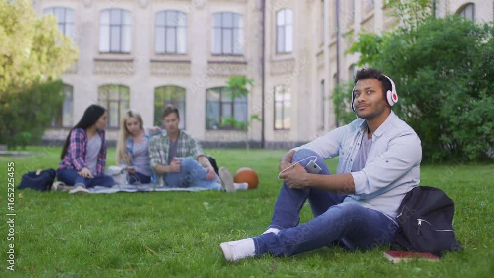 Handsome Hispanic student sitting on grass and enjoying music in headphones Handsome Hispanic student sitting on grass and enjoying music in headphones
