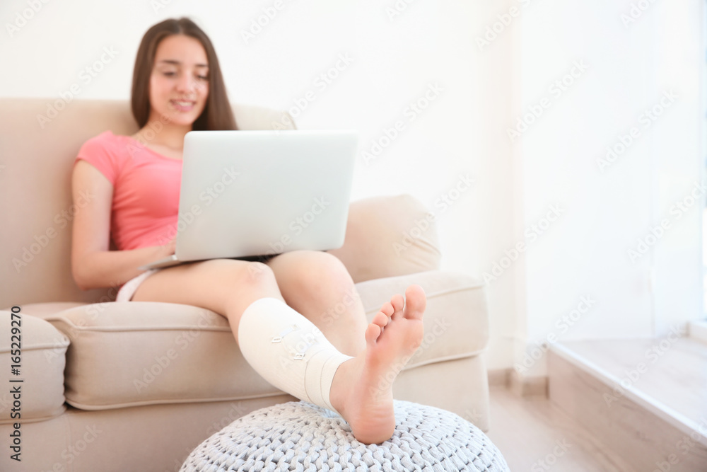 Beautiful young woman with laptop and bandaged leg sitting on sofa at home Beautiful young woman with laptop and bandaged leg sitting on sofa at home