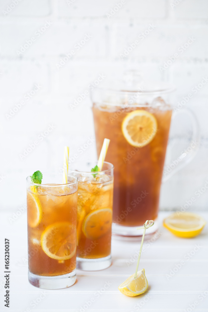 Two Glasses and Pitcher Filled with Iced Tea Ice Cubes and Lemon Slices Garnished with Mint and Striped Straws Against the White Brick Wall on the Wooden Table