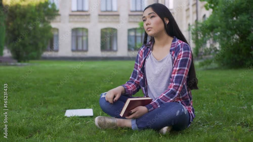 Thoughtful Asian female student sitting on lawn alone and missing parents Thoughtful Asian female student sitting on lawn alone and missing parents
