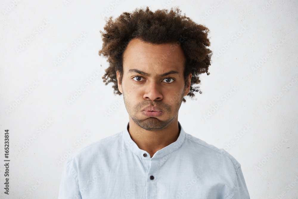 Fototapeta premium Isolated studio shot of unhappy grumpy serious young African American manager with Afro hairstyle frowning, puffing his cheeks, feeling fed up as he has to stay at office and work extra hours.