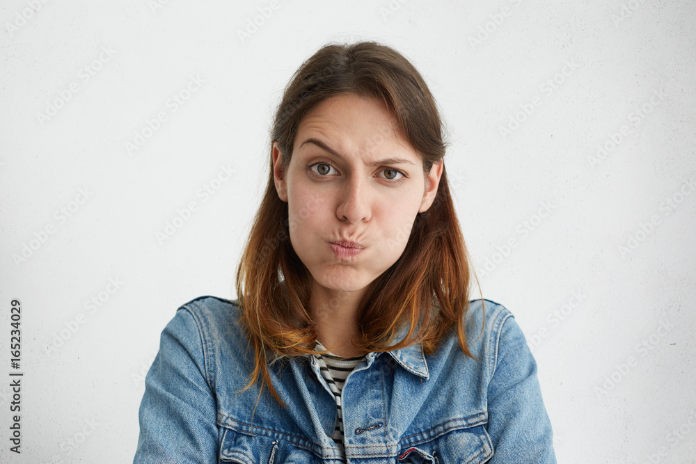 Headshot of displeased European woman blowing her cheeks being upset raising her eyebrows, demonstrating her discontent. Female in jean jacket with sulky expression isolated over white background Headshot of displeased European woman blowing her cheeks being upset raising her eyebrows, demonstrating her discontent. Female in jean jacket with sulky expression isolated over white background