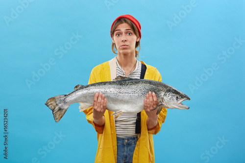 Studio portrait of beautiful female wearing red hat, yellow raincoat and jean overalls frowning her face while holding huge fish in hands. Successful angler with her catch. Hobby, recreation concept