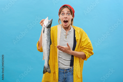Human facial expressions, emotions and feelings concept. Young attractive female with surprised expression holding huge fish, surprised with successful catch posing against studio wall background.