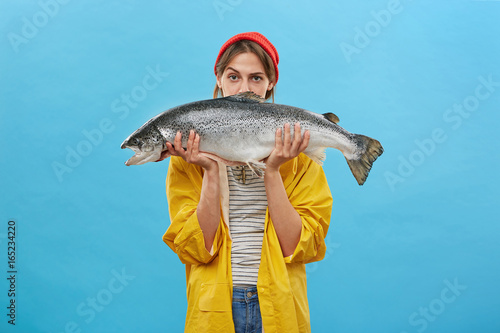 Pretty housewife holding huge fish caught by her husband. Beautiful woman dressed in yellow raincoat and red hat demonstrating her successful catch isolated over blue backgound. Fishery and fishing