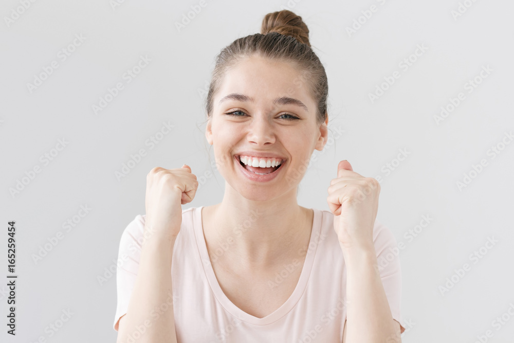 Indoor photo of good-looking teenage girl isolated on gray background celebrating victory and acting as if she is winner, squeezing fists in deep emotional expression of happiness, luck and content. Indoor photo of good-looking teenage girl isolated on gray background celebrating victory and acting as if she is winner, squeezing fists in deep emotional expression of happiness, luck and content.