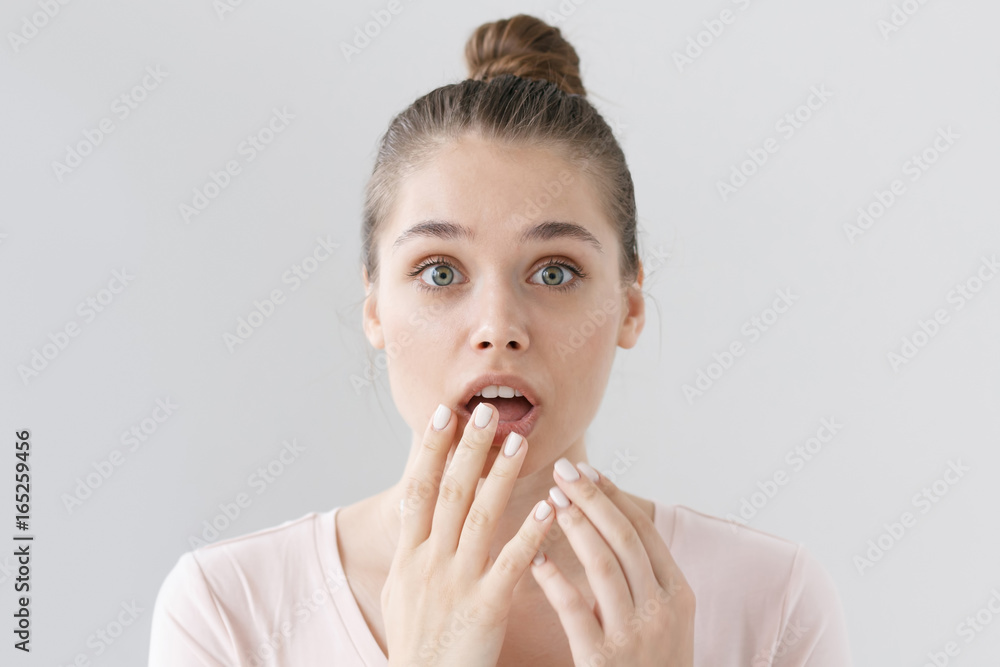 Horizontal headshot of frightened young woman with hair tied in bun isolated on grey background with eyes round because of astonishment and surprise, trying to cover round mouth with both hands. Horizontal headshot of frightened young woman with hair tied in bun isolated on grey background with eyes round because of astonishment and surprise, trying to cover round mouth with both hands.