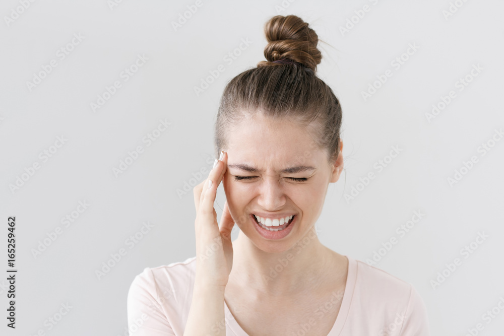 Horizontal indoor shot of beautiful young woman isolated on gray background squeezing face because of strong pain, putting one hand to head, screaming and curving mouth in suffering from migraine. Horizontal indoor shot of beautiful young woman isolated on gray background squeezing face because of strong pain, putting one hand to head, screaming and curving mouth in suffering from migraine.