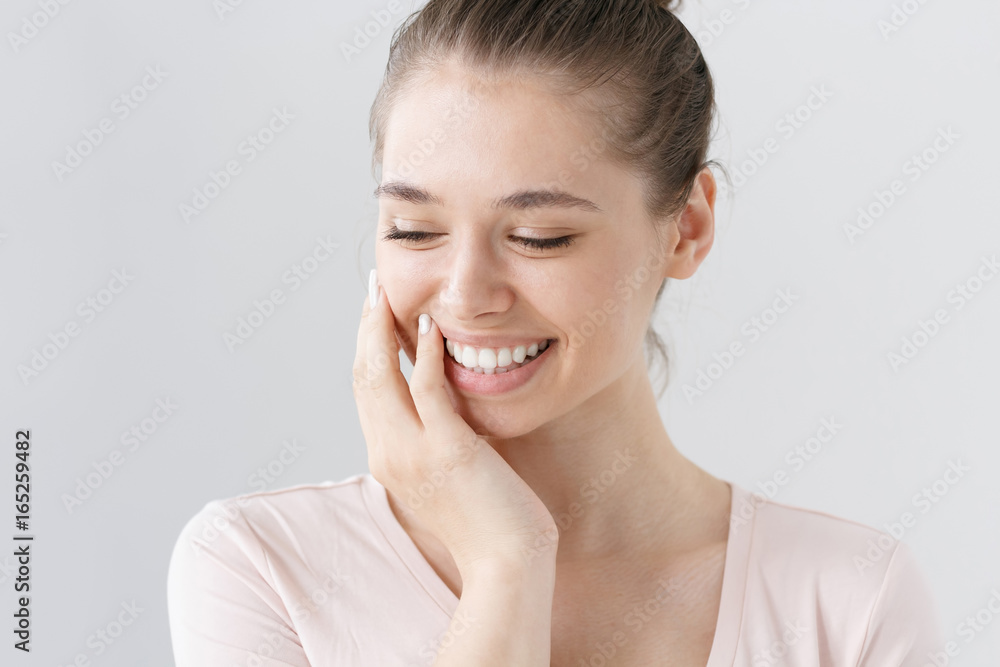 Indoor portrait of naturally beautiful young woman isolated on gray background with eyes looking down, being shy, touching cheek with hand in gesture of uncertainty and secrecy, looking feminine. Indoor portrait of naturally beautiful young woman isolated on gray background with eyes looking down, being shy, touching cheek with hand in gesture of uncertainty and secrecy, looking feminine.