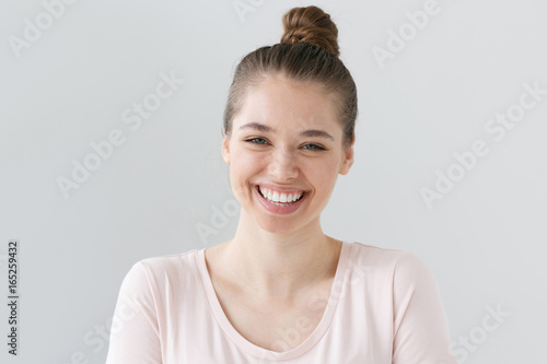 Closeup of positive teenage girl with brown hair tied in bun isolated on gray background with expression of happiness on face  laughing emotionally with excitement as if reacting to funny joke.