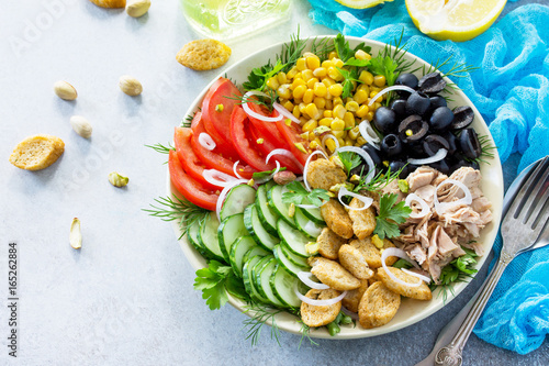 Salad with tuna, fresh tomato, cucumber, onion, pickled olives, corn and salad dressing vinaigrette on a gray stone background or slate. Copy space.