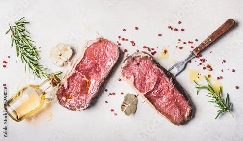 Raw beef Striploin steaks with oil , spices and meat fork on white stone background, top view, flat lay, horizontal