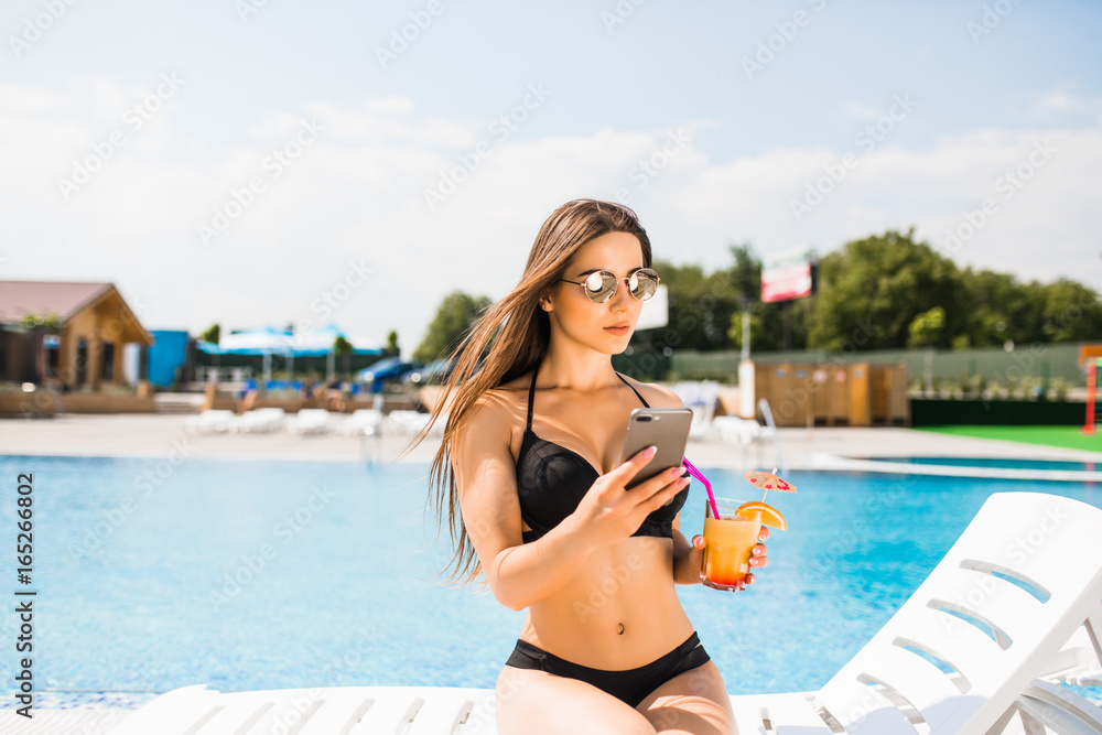 Young woman in swimsuit relaxing with cocktail near pool and use phone