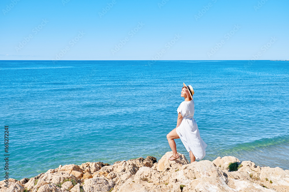 Beautiful girl in a summer dress and hat on the seashore near a background old city europe. Mediterranean Sea, Sitges, Spain Beautiful girl in a summer dress and hat on the seashore near a background old city europe. Mediterranean Sea, Sitges, Spain