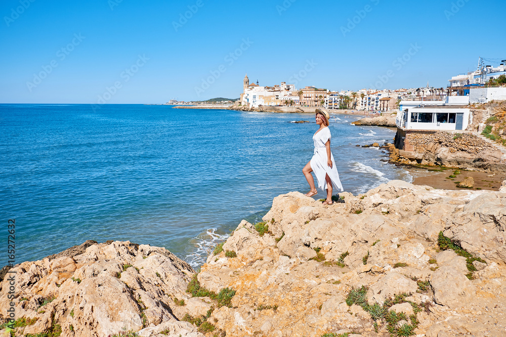Beautiful girl in a summer dress and hat on the seashore near a background old city europe. Mediterranean Sea, Sitges, Spain Beautiful girl in a summer dress and hat on the seashore near a background old city europe. Mediterranean Sea, Sitges, Spain