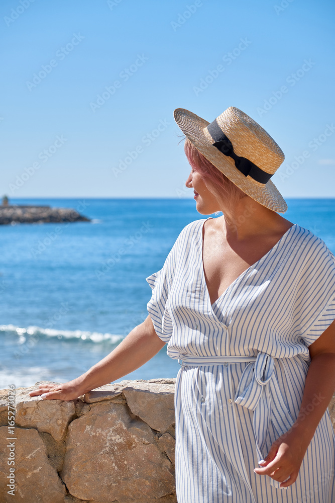 Beautiful girl in a summer dress and hat on the seashore near a background old city europe. Mediterranean Sea, Sitges, Spain