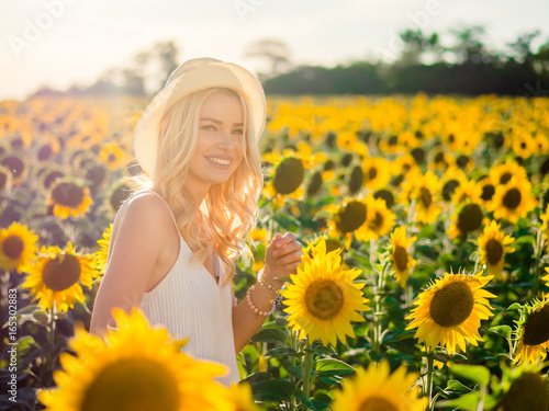 Young beautiful blonde woman standing in sunflower field. Sunset background. Sexy sensual portrait of girl in straw hat and white summer dress.