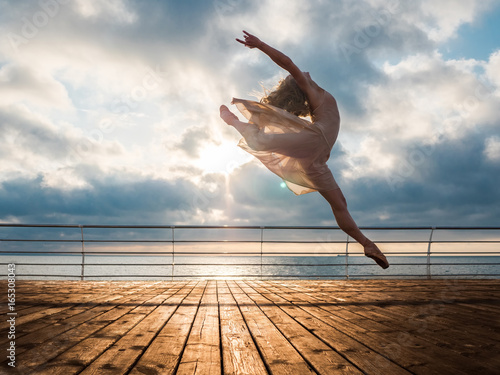 Fotografia, Obraz Jumping ballerina in beige dress and pointe on embankment above ocean or sea beach at sunrise