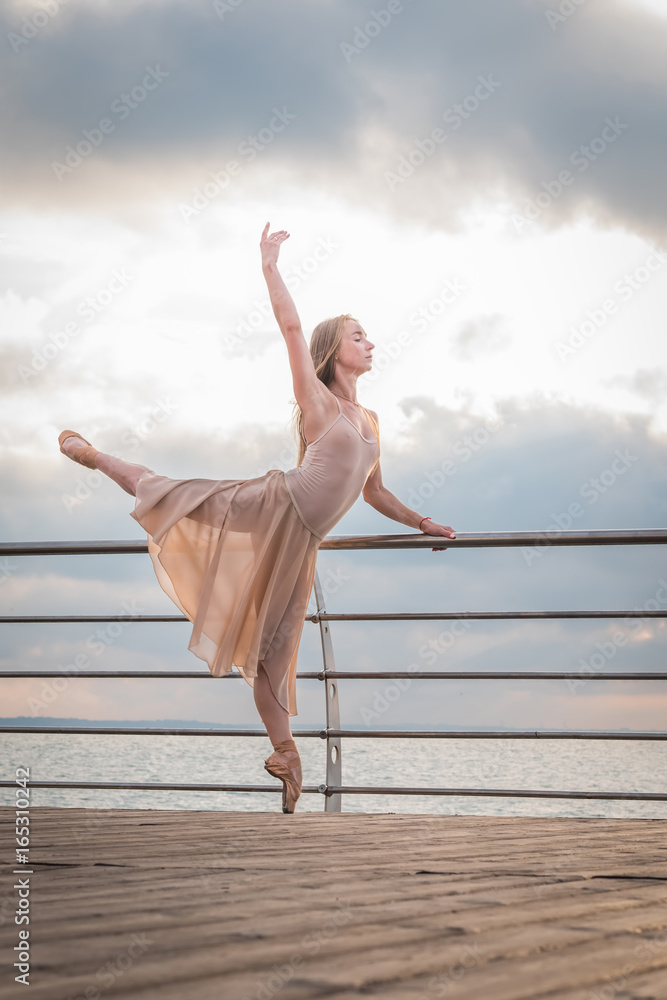Dancing ballerina in beige silk dress and pointe on embankment above ocean or sea beach at sunrise. Young beautiful blonde woman with long hair practicing classic exercises with emotions.