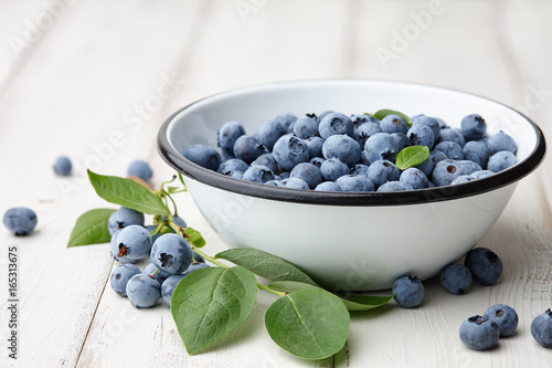 Fresh ripe blueberries with leaves in bowl on white wooden planks